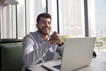Cheerful handsome Arab online project executive man sitting at laptop, working business tasks at co-working table, looking away, smiling, laughing, thinking on successful marketing strategy