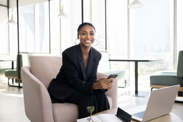 Confident beautiful young African entrepreneur woman looking at camera with happy smile, sitting in armchair, working in comfortable co-working space, holding mobile phone. Business portrait