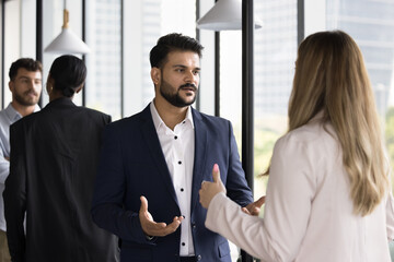 Serious young Indian businessman talking to Caucasian female colleague in office hall at short meeting. Diverse co-workers discussing business strategy, planning project communication