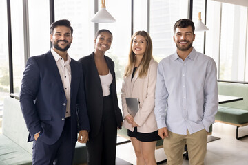 Happy multiethnic team of young business colleagues standing for group portrait in office co-working space, looking at camera, smiling, promoting diversity, corporate success, teamwork
