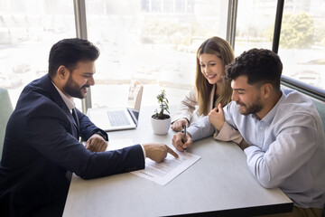 Happy young Indian realtor man pointing finger at document for cheerful husband and wife signing, giving legal consultation, support. Positive couple setting signature in loan agreement
