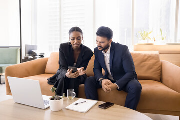 Young African businesswoman showing online content on smartphone to Indian colleague man, discussing project presentation, application for job, sitting on couch in co-working office