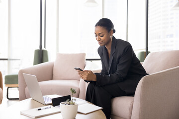 Confident beautiful African American businesswoman using cellphone for job communication, reading online content, typing, chatting on mobile phone, sitting in armchair in co-working office