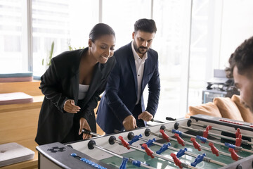 Positive excited young multiethnic couple of professionals playing toy football, competing in table soccer with coworkers, turning handles at field miniature, laughing, relaxing on work break