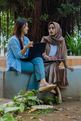 A pair of elegant candid Asian girl discussing studying together holding laptop, books and clipboard in campus park. female students for education, promotion and lifestyle concepts