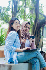 two cheerful indonesia women to the camera showing off online transaction debit cards by holding pens, books and clipboards in the campus park. students for education and lifestyle concepts