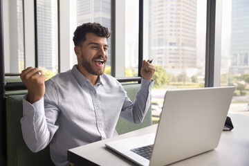 Happy joyful Arab business man enjoying win, victory, achieving job success, making winner yes hands gesture at laptop, sitting at co-working workplace table, shouting, getting profit