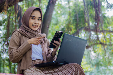 Beautiful Asian woman in hijab sitting smiling at the camera pointing at her smartphone gadget in front of her laptop in the park. female student for education, technology and lifestyle concept