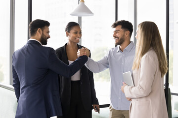 Cheerful young diverse business colleagues shaking hands in office hall, smiling, laughing, celebrating successful startup, congratulating each other on job success, giving friends handshake gesture