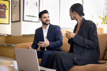 Diverse business colleagues wearing formal suits talking at laptop on couch. Professional man and woman discussing work project on break, drinking tea, meeting at computer in coworking space