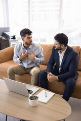 Serious young Arab employee man talking to Indian colleague, moving hands for explaining. Multiethnic business coworkers meeting in office co-working hall, sitting on couch, Vertical shot