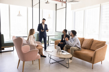 Multiethnic small business team and Indian male boss meeting in modern co-working space, sitting on soft couch, listening to speaking leader, discussing project strategy