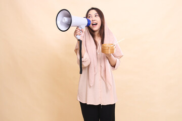 Asian woman wearing a hijab looks to the left shouting loudly angrily carrying a megaphone (loudspeaker) to make announcements and holding a wooden bowl containing delicious dim sum