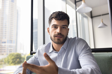Serious young business professional man talking on video call, sitting in co-working cafe, looking at camera, speaking, giving consultation on online conference chat. Head shot portrait, screen view