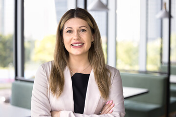 Happy Caucasian professional woman posing in co-working space, looking at camera, standing with hands folded, smiling. Positive legal adviser, business consultant, CEO, executive portrait