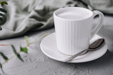 White cup of frothy milk coffee on a textured table with a spoon and green leaves