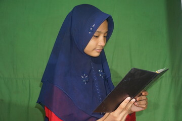 A teenage girl in a hijab is reading a book on a green background. Southeast Asian Teenager