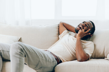 Happy African American Man Talking on Black Smartphone while Sitting on a Modern White Sofa at Home...