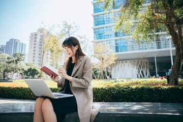 Young Asian woman looking up while using laptop, sitting at park against office buildings in the...