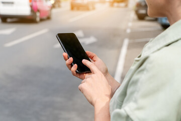 Modern female person stands next to road in city and using her smartphone. Searching car rental, taxi, public transportation.