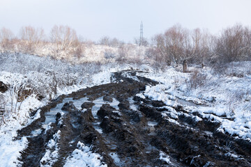 Tire tracks on muddy road with iced puddles, snowy landscape in winter