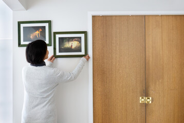 Woman hanging picture frames on a white wall at home. Fairytale glowing mushroom photos in the frames. - 789985362