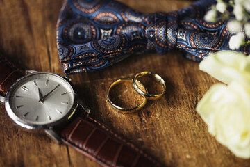 Golden wedding rings. Rustic wooden table. Buttonhole flowers. Wedding accessories background. Man fashion. Elagant fabric bowtie. Leather watch strap. Time to get married.