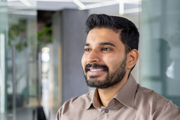 Obraz premium A portrait of a confident young man with a beard smiling warmly in a well-lit, contemporary office environment. The image conveys a sense of happiness and professional success.
