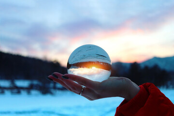Closeup womans hand holding crystal sphere with scenic cloudy sunset on the background. Winter in...