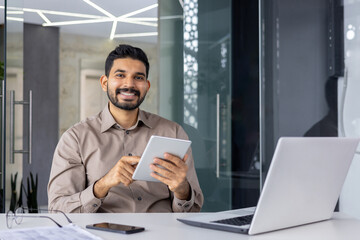 Fototapeta premium A confident young businessman with a warm smile works on a tablet while sitting at a desk in a sleek, contemporary office environment. He displays professionalism and approachability.