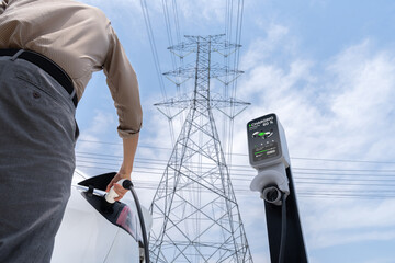 Man recharge EV electric car battery at charging station connected to electrical power grid tower...