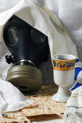 Gasmask, partially concealed by white prayer talit, next to a Kaddish cup on a table with matzah...