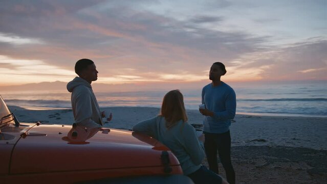Young couple with friends on vacation standing by car at beach watching morning sunrise and drinking coffee on road trip  - shot in real time