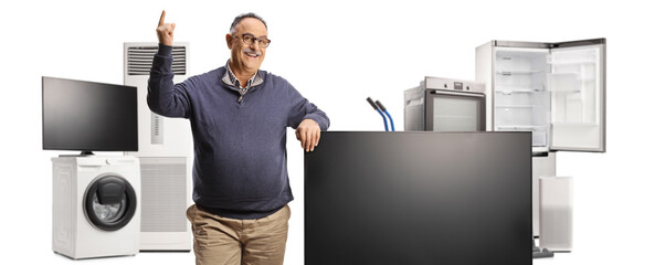 Mature man pointing up and standing next to a flat tv screen and other home electric appliances - obrazy, fototapety, plakaty