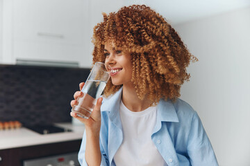 Young African American woman enjoying a refreshing drink of water in her stylish kitchen at home - obrazy, fototapety, plakaty