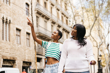 A young female couple is exploring a new city during a short weekend trip, walking together while pulling a suitcase along a city street.