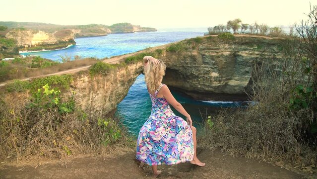 a woman in a floral chiton stands on a cliff overlooking the ocean