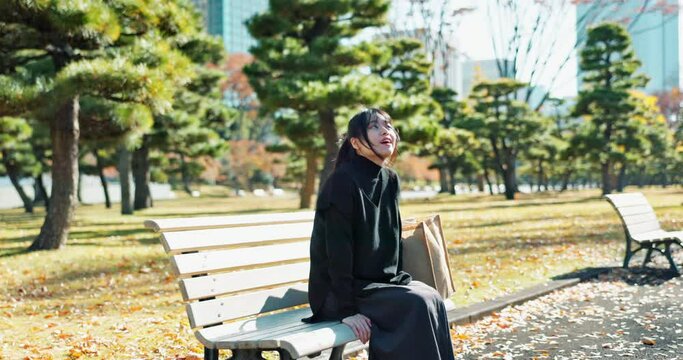 Japanese woman, bench and smartphone in park with relax, happiness and social media in sunshine in city. Female person, sitting and wellness with cellphone in town, streaming and digital technology
