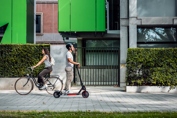 Cheerful woman riding a bicycle crosses a concentrated man on an electric scooter through the city - 790066128
