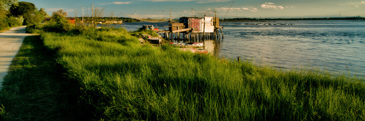 Tranquil waters embrace a picturesque scene of fishing boats and rustic huts along the shores of Pialassa dei Piomboni, near Ravenna - obrazy, fototapety, plakaty