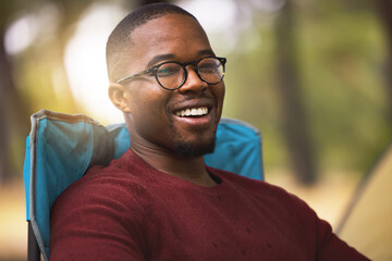 Camping, portrait and black man relax outdoor on vacation in wilderness for peace and wellness in Norway. Person, happy and chair in forest woods for adventure in nature, summer holiday and fresh air
