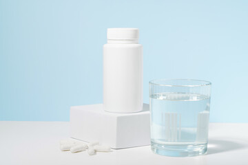White mockup of a pill jar on a pedestal and a glass of water on a blue background. Concept of medicine, pharmacy, vitamins for beauty and health, dietary supplements, and treatment of diseases.