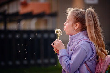 A young girl in a lilac jacket captures the whimsy of childhood as she blows on a dandelion, with...