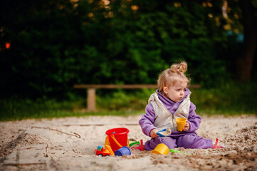 Engrossed in play, a young toddler fills a cup with sand, surrounded by colorful toys in a sandbox, a picture of focus and discovery