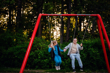 An adult man and woman enjoy swinging on a playground at twilight, their smiles reflecting carefree happiness amidst a backdrop of lush greenery.