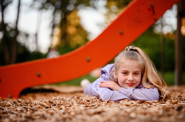 A young girl rests her head in her hands on the playground woodchips, her thoughtful gaze and the...