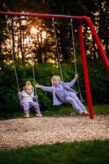 Two children enjoy a serene sunset as they swing in a playground surrounded by lush greenery, encapsulating the joy and innocence of childhood