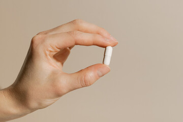 Female hand holding white pill capsule with fingers on beige isolated background. Concept of pharmacy, medicine, dietary supplements, vitamins and depression treatment.