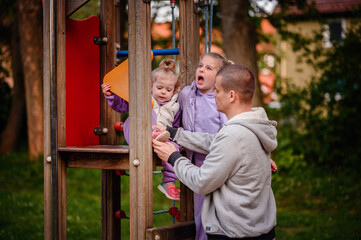 A playful day unfolds as dad helps put on his youngest daughter's shoes while the second daughter makes faces, having fun, highlighting family fun and outdoor activities