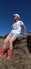 Closeup photograph of a Caucasian female with her hair in  a plat, sitting on a round bale of hay on a sunny day wearing shorts, t-shirt a cap and pink boots, looking up into the blue sky